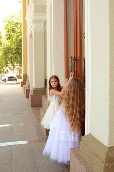 Две очаровательные маленькие девочки в длинных белых платьях стоят возле зеркальных дверей здания на открытом воздухе — стоковое фото