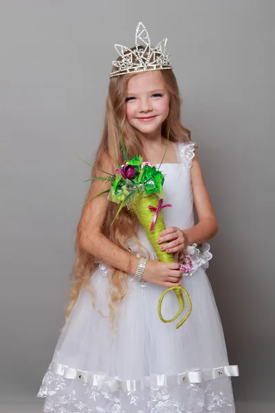 Caucasian little girl with beautiful long hair in the crown and a white dress with a cute smile posing at the camera on a gray background — Stock Photo, Image