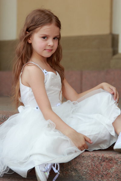 Portrait of cute smiling little girl with long hair in a beautiful dress is sitting on the steps outdoors