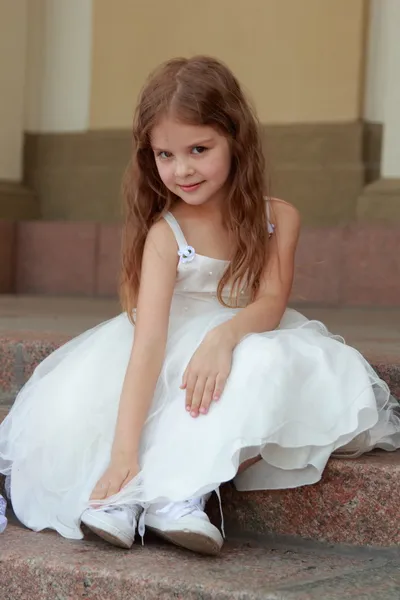 Smiling cheerful little girl in a beautiful white ball gown and sneakers sitting on the stairs to the outdoors — Stock Photo, Image