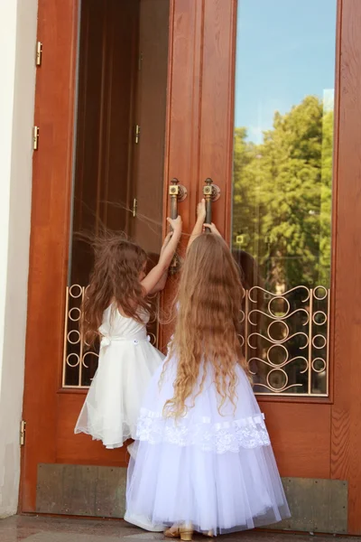 लंबी सफेद पोशाक में दो आकर्षक छोटी लड़कियों इमारत के दर्पण दरवाजे के पास खड़े हैं — स्टॉक फ़ोटो, इमेज