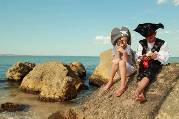 Boy and girl in a pirate costume with a map and a magnifying glass sitting on a large rock by the sea — Stock Photo, Image