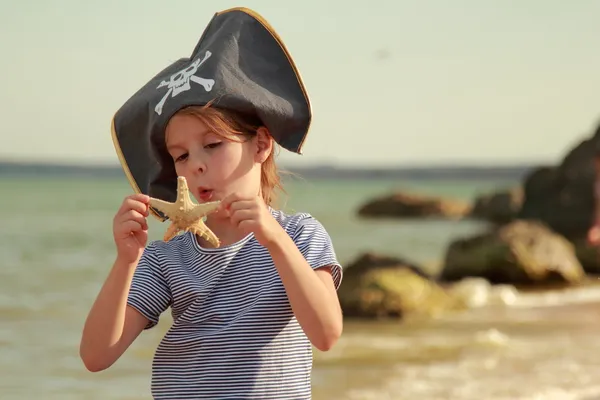 Schönes kleines Mädchen mit Piratenmütze und Totenkopf, das einen Seestern am Strand hält — Stockfoto