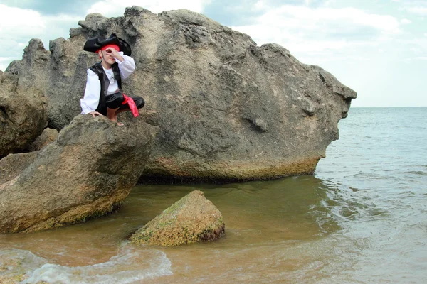 Mosolygó fiatal fiú öltözött, mint egy kalóz, keres egy távcső, ül egy nagy szikla a tengerparton — Stock Fotó