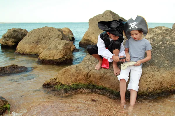 Junge und Mädchen im Piratenkostüm mit Landkarte und Lupe sitzen auf einem großen Felsen am Meer — Stockfoto