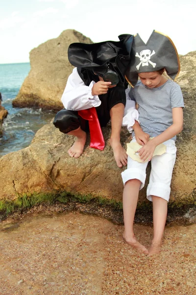 Garçon et fille en costume de pirate avec une carte et une loupe assis sur un grand rocher au bord de la mer — Photo