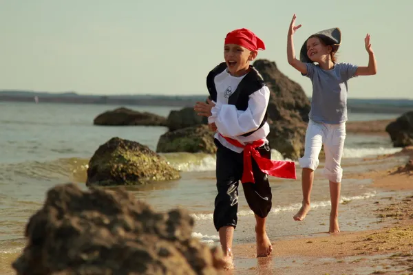 Fröhlich lächelnde kleine Jungen und Mädchen in Piratenkostümen laufen an heißen Sommertagen barfuß am Meer entlang — Stockfoto
