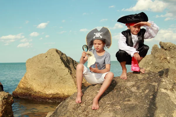 Boy and girl in a pirate costume with a map and a magnifying glass sitting on a large rock by the sea — Stock Photo, Image