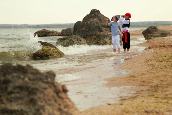 Fröhlich lächelnde kleine Jungen und Mädchen in Piratenkostümen laufen an heißen Sommertagen barfuß am Meer entlang — Stockfoto