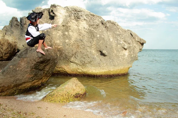 Sorridente giovane ragazzo vestito da pirata guardando attraverso un telescopio seduto su una grande roccia al mare — Foto Stock