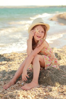 Smiling young girl holding a starfish sitting on the bank of the summer sea.