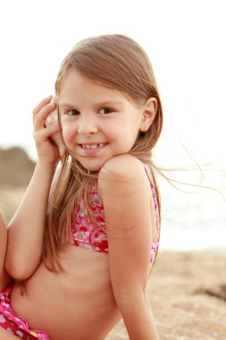 sevimli küçük kız bir mayo içinde gülümseyen deniz kum bir arka plan üzerinde oturur.