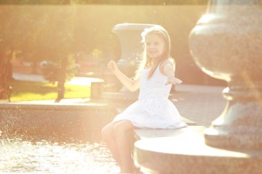 Charming cheerful little girl in a white dress and enjoying the cool water splashing in the fountain clipart
