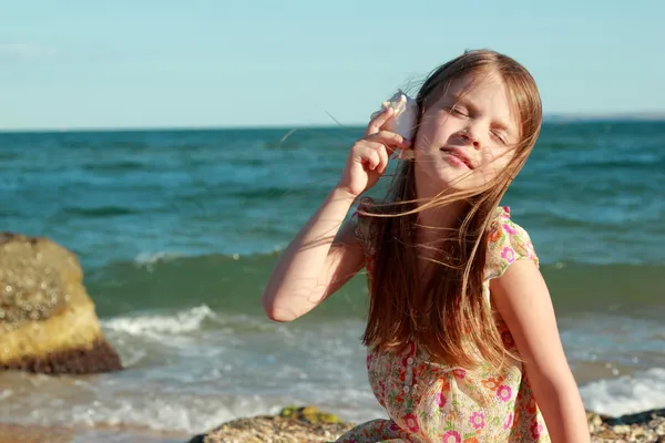 Portret van een schattig klein meisje met dromen van een glimlach en houden een zeeschelp zit op de bank van de zomer zee. — Stockfoto
