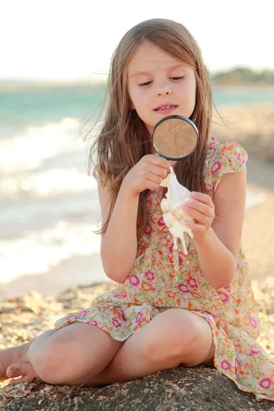Blije jonge meisje onderzoekt een vergrootglas zeeschelp op de strand zomer zee — Stockfoto