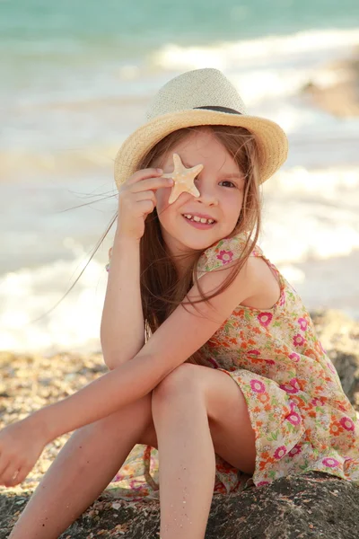 Portrait of a cute little girl with dreams of a smile and holding a seashell sits on the bank of the summer sea. — Stock Photo, Image
