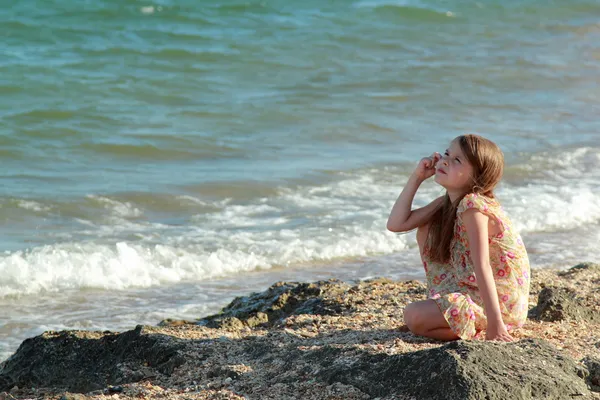 Retrato de uma menina bonito com sonhos de um sorriso e segurando uma concha senta-se na margem do mar de verão . — Fotografia de Stock