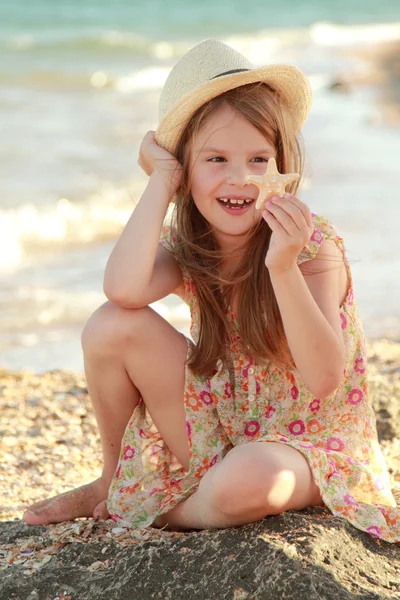 Portret van een schattig klein meisje met dromen van een glimlach en houden een zeeschelp zit op de bank van de zomer zee. — Stockfoto