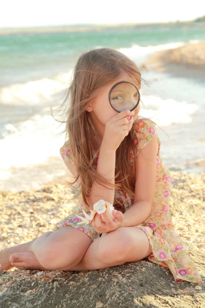 Glada unga flickan undersöker en förstoringsglas snäcka på stranden sommaren havet — Stockfoto