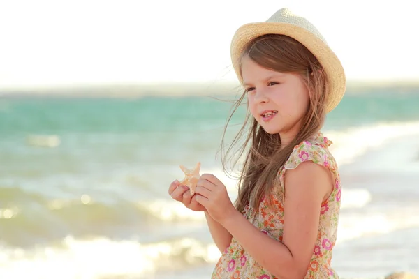 Caucasian cute young girl in a summer dress and hat holding a starfish — Stock Photo, Image