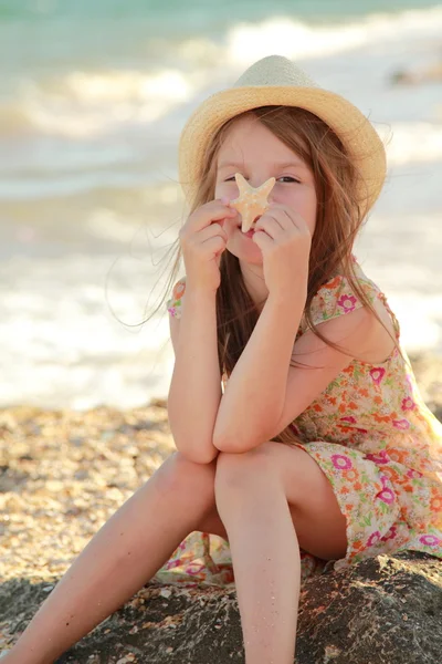 Sorrindo menina segurando uma estrela do mar sentado na margem do mar de verão . — Fotografia de Stock