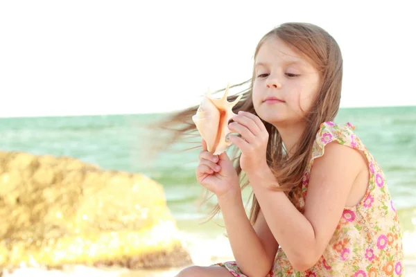 Adorable happy little girl holding a seashell on the sea beach on a sunny day. — Stock Photo, Image