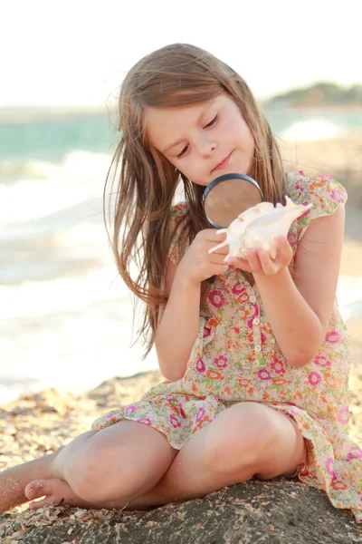 Joyful young girl examines a magnifying glass seashell on the beach summer sea. — Stock Photo, Image