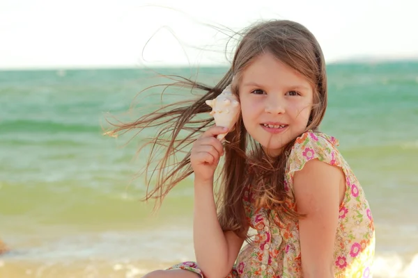 Retrato de una linda niña con sueños de una sonrisa y sosteniendo una concha de mar se sienta en la orilla del mar de verano . — Foto de Stock