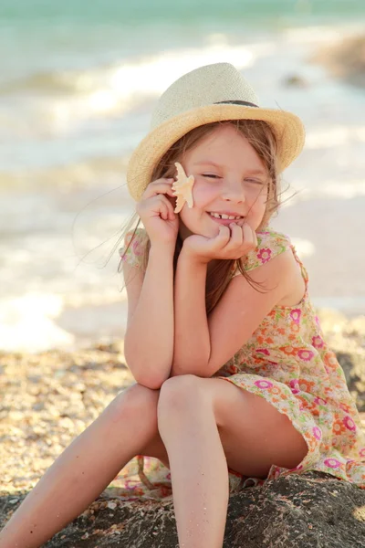 Sorrindo menina segurando uma estrela do mar sentado na margem do mar de verão . — Fotografia de Stock