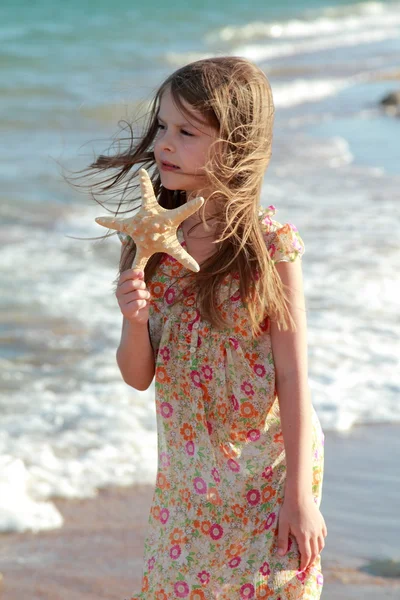 Entzückend glückliches kleines Mädchen hält einen Seestern am Strand in den sonnigen Tag. — Stockfoto