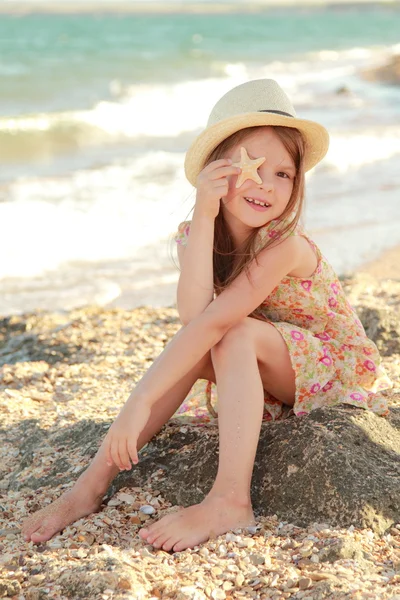 Sorrindo menina segurando uma estrela do mar sentado na margem do mar de verão . — Fotografia de Stock