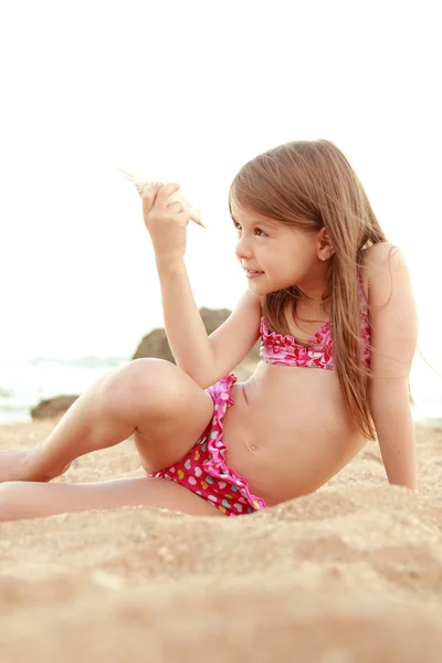 Härlig liten flicka i en baddräkt som leker i sanden med en snäcka. — Stockfoto