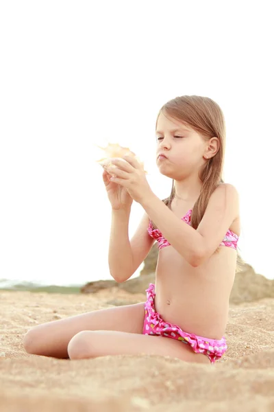 Beautiful cheerful young girl sitting on the sand and holds a large seashell. — Stock Photo, Image