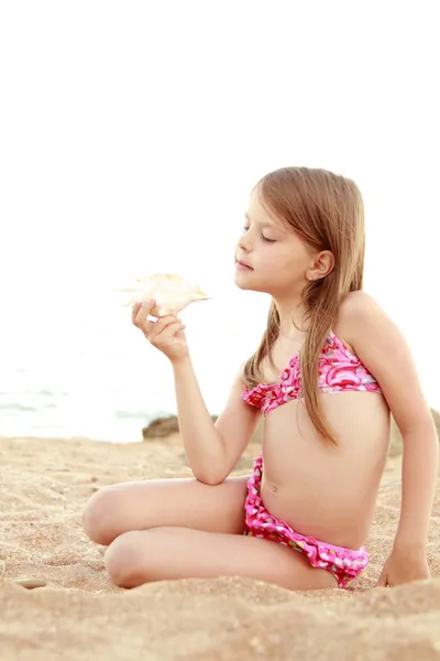 Menina adorável em um maiô brincando na areia com uma concha . — Fotografia de Stock