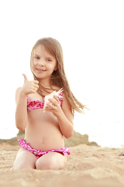 Schattig gelukkig meisje met een charmante glimlach spelen in het zand met een zeeschelp. — Stockfoto