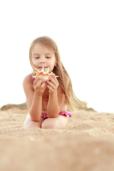 Portrait of a smiling pretty little girl in a swimsuit holding a beautiful seashell. — Stock Photo, Image