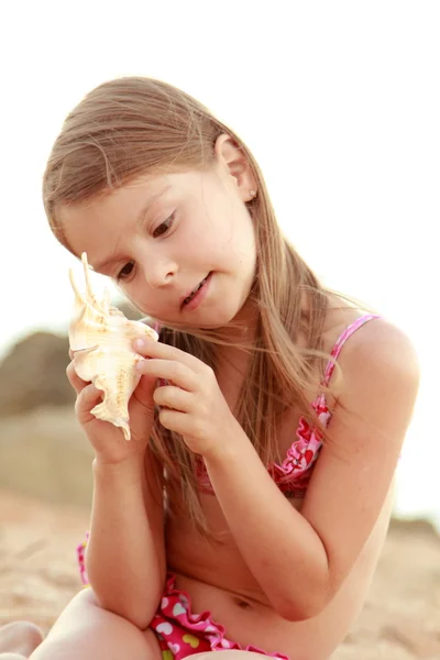 Caucasian happy young girl in a pink swimsuit sitting on the beach and played with a seashell. — Stock Photo, Image