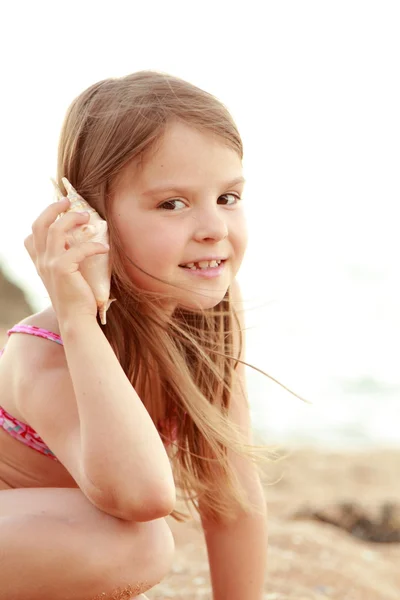 Sevimli küçük kız bir mayo içinde gülümseyen deniz kum bir arka plan üzerinde oturur. — Stok fotoğraf