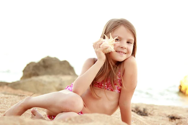 Söt glad tjej med ett charmigt leende leker i sanden med en snäcka på stranden. — Stockfoto