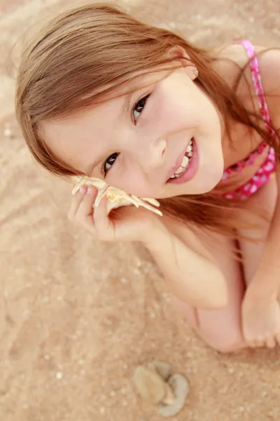 Beautiful cheerful young girl sitting on the sand and holds a large seashell. — Stock Photo, Image