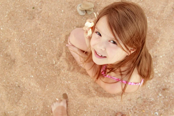 European smiling little girl in a pink swimsuit sitting on a background of sea sand. — Stock Photo, Image