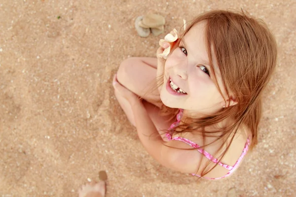 Europeo sorridente bambina in costume da bagno rosa seduta su uno sfondo di sabbia marina — Foto Stock