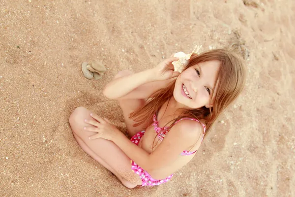 Kum üzerine oturan neşeli güzel genç kız ve büyük bir deniz kabuğu tutar. — Stok fotoğraf