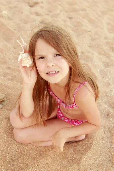 Mooie vrolijke jonge meisje, zittend op het zand en bezit een grote zeeschelp. — Stockfoto