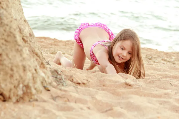 Hermosa niña sonriente en un traje de baño rosa se juega en la arena en la playa . — Foto de Stock