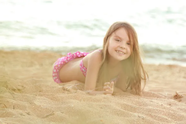 Bela menina sorridente em um maiô rosa é jogado na areia na praia . — Fotografia de Stock