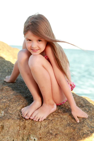 Linda niña en un traje de baño sentado en una gran roca junto al mar . — Foto de Stock