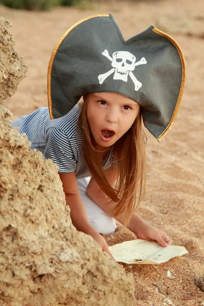 Carino ragazza in un cappello da pirata con una mappa dei pirati in mano su uno sfondo . — Foto Stock