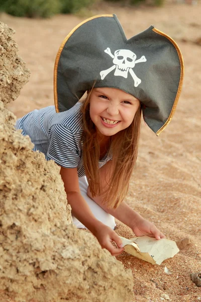 Menina bonito em um chapéu de pirata com um mapa pirata na mão em um fundo de areia do mar . — Fotografia de Stock