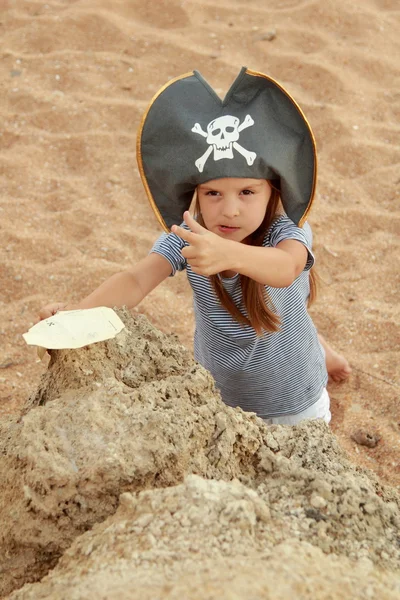 Cute young girl in a pirate hat with a pirate map in hand on a background of sea sand on the beach. — Stock Photo, Image